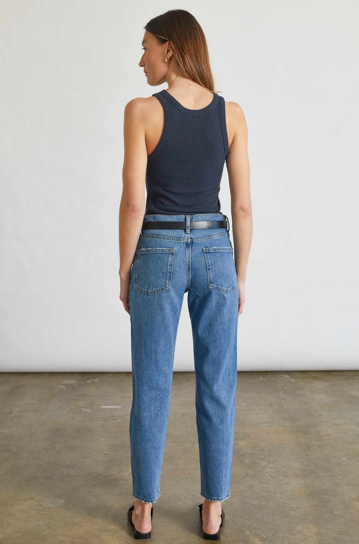 Woman wearing a navy tank top and blue jeans standing against a plain white wall.