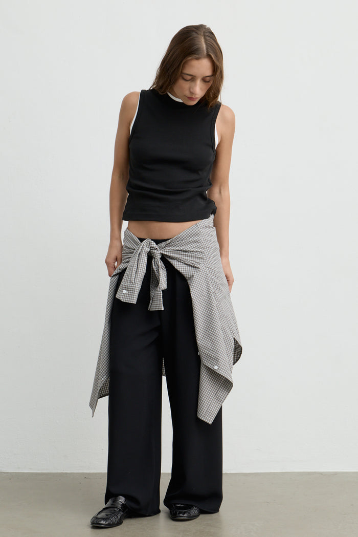 A woman stands against a plain light gray wall wearing a black sleeveless mock-neck top, black wide-leg pants, and a black-and-white patterned shirt tied around her waist.