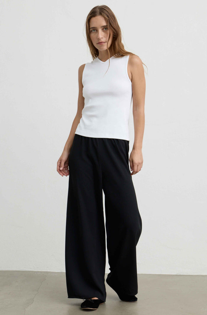 A woman modeling a sleeveless white mock-neck top paired with wide-leg black trousers, standing against a plain light gray studio background.
