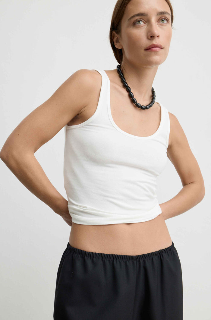 A woman wearing a white scoop-neck tank top and black pants, standing against a light gray studio background.