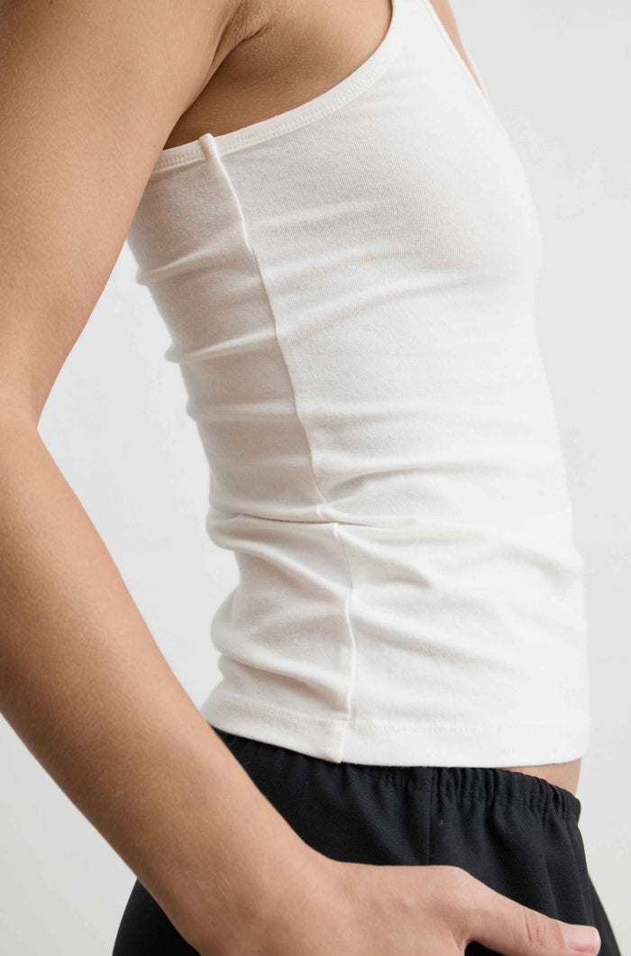 Close-up of a person wearing a fitted white tank top and black shorts against a light gray studio background.