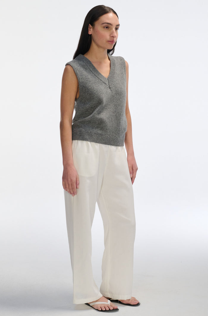 Woman wearing a gray sleeveless sweater and white pants on a white background